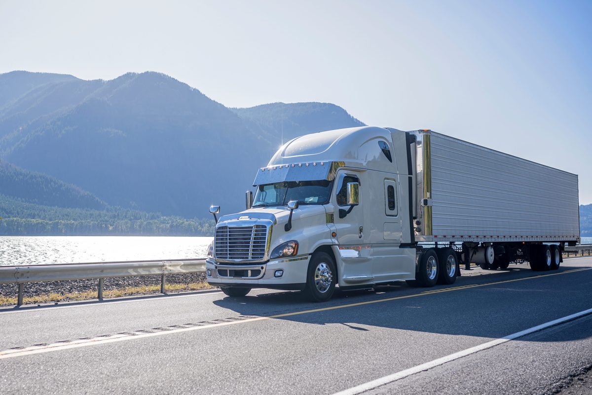 White semi-truck hauling a refrigerated trailer on a highway beside a lake and mountains